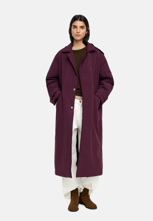 Kobieta w długim, ciemnofioletowym pikowanym płaszczu, brązowym swetrze, białych szerokich spodniach i brązowych botkach za kostkę, stojąca z rękami w kieszeniach.