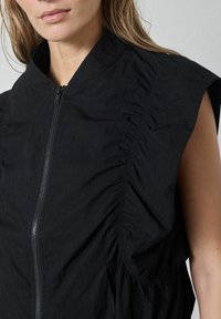 Čierny bezrukávový odev s vysokým golierom, stredným zipsom a nariasenou textúrou pozdĺž prednej a bočných strán. Hladký povrch látky.