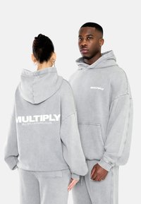 Grijze hoodies met grote "MULTIPLY" tekst op de achterkant en een kleiner logo op de voorkant. Zachte textuur, oversized pasvorm en kangoeroe pockets.