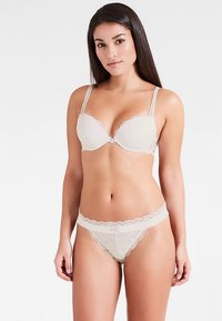 Reggiseno color beige in pizzo con bordi smerlati e spalline regolabili, abbinato a un perizoma in pizzo coordinato che mostra un delicato motivo floreale.