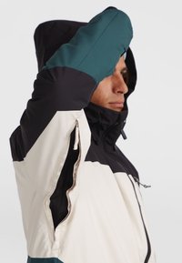 Geïsoleerde jas met een zwarte capuchon, teal mouwen en een crèmekleurig lichaam. Voorzien van ritsventilatieopeningen en een hoge kraag voor extra bescherming.