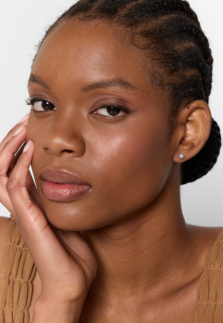 Nahaufnahme einer Person mit geflochtenem Haar, die Ohrstecker trägt. Das Model hat glatte Haut und trägt ein Oberteil in neutralen Tönen aus strukturiertem Stoff.