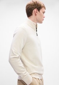 Pulover cu fermoar la jumătate de lungime, de culoare bej deschis, cu textură ribbed pe umeri și fermoar negru. Material moale și design ajustat, cu mâneci lungi.