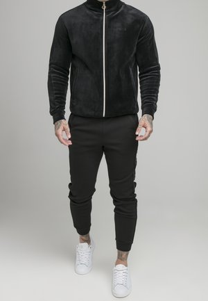 Mand iført sort lynlåstrøje, sorte joggers og hvide sneakers, stående mod en lysegrå baggrund.
