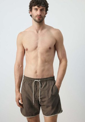 Man met krullend donker haar, gekleed in bruine zwemshorts met witte koordsluiting, staand tegen een effen lichte achtergrond.