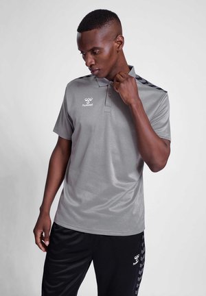 Mężczyzna w szarej koszulce polo i czarnych spodniach z logo Hummel, poprawiający kołnierz koszuli, stojący na jednolitym jasnym tle.