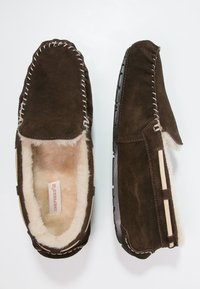 Pantofole in suede marrone con una morbida fodera in pelliccia, dettagli cuciti a mano e suola in gomma piatta. Accentuate con strisce di pelle chiara.