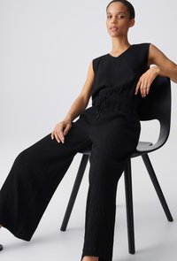 Femme assise sur une chaise noire, portant une combinaison noire texturée sans manches avec une ceinture à la taille, sur un fond clair uni.