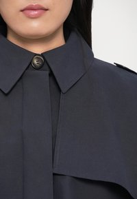 Trenci bleumarin cu un guler mare, detalii cu nasturi și o textură netedă, având un buzunar lateral și un design curat, minimalist.