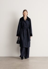 Ναυτικό παλτό τραντσκότ με ζώνη στη μέση, φαρδιά γιακά και μεγάλες τσέπες. Το μοντέλο φοράει παντελόνια με φαρδιά πόδια και κρατάει μια μικρή μαύρη τσάντα χειρός.