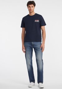 Navyblauwe katoenen t-shirt met korte mouwen, met een witte "JEANS" graphic op de linkerkant van de borst. Gecombineerd met lichtblauwe denim jeans en witte sneakers.