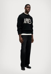 Zwarte gebreide trui met 'APRÈS' in het crèmekleurig, gecombineerd met donkere spijkerbroek en zwarte schoenen. De pasvorm is los, met geribbelde boorden.