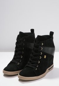 Svartsko med kilklackar och beige sula, snörning fram och guldlogotyp på sidan, visade mot en enkel bakgrund.