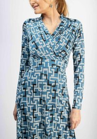 Pitkähihaisen sinisen kankaan mekko, jossa on geometrinen mustavalkoinen kuvio, kietoutumismalli ja sileä rakenne.