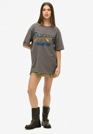 Grå oversized t-shirt med et grafisk design med teksten "Supercar North American Tour '79" i blå og guld, parret med khaki shorts og sorte støvler.