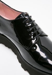 Chaussure en cuir verni noir avec un bout arrondi, un design à lacets, une semelle noire texturée et des détails de couture minimalistes.