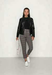 Juodas dirbtinės odos švarkas su užtrauktuku priekyje, derinamas su raštiniais kelnėmis rudos ir kreminės spalvos atspalviuose, ir baltais sportbačiais. Laiko baltą rankinę.