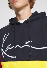 Felpa con cappuccio con base nera e sezioni rosse e gialle, logo ricamato bianco, coulisse e tessuto morbido al tatto.