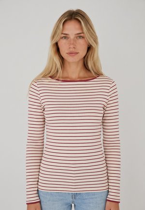 Mujer con cabello largo y rubio que lleva una camisa blanca de manga larga con finas rayas horizontales rojas y vaqueros azul claro, de pie sobre un fondo liso.