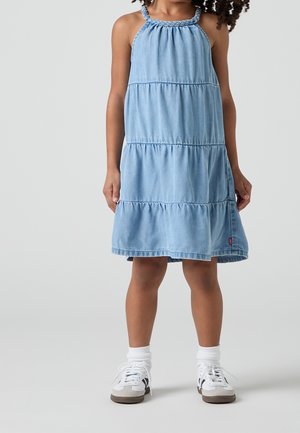 Niño con vestido azul claro de capas sin mangas y escote trenzado, calcetines blancos y zapatillas blancas, de pie sobre un fondo liso.