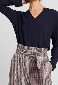 Nainen, jolla on yllään tummansininen V-pääntie pitkähihaisten yläosa, joka on sisätaskuissa korkeavyötäröisten ruutukuosisien housujen sisällä, ja hänellä on käsi taskussa.