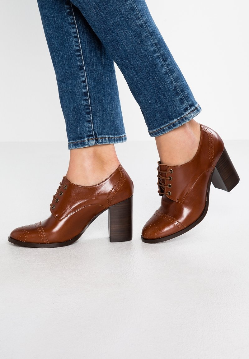 Chaussures à talons en cuir marron avec un embout arrondi, un laçage à l'avant et des détails de brogue. Le talon est de forme bloc avec une finition en bois plus foncé.
