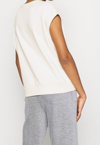 Top sin mangas de color beige claro con escote redondeado, ajuste relajado y dobladillo acanalado, combinado con pantalones de chándal grises que presentan una textura lisa.