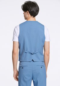 Ljusblå skräddarsydd väst med en slät yta, bakre knyt-detalj och en något figursydd form. Kombinerad med en vit t-shirt under.