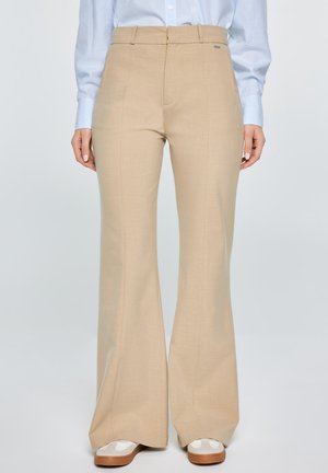 Beżowe, wysokie spodnie z szerokimi nogawkami o subtelnej fakturze, z bocznymi kieszeniami i metalowym akcentem, zestawione z białymi trampkami i jasnoniebieską koszulą.