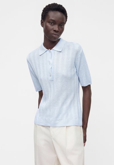 Femme portant un polo texturé bleu clair à manches courtes et un pantalon ample blanc cassé, se tenant devant un fond blanc uni.