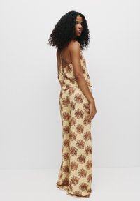 Mouwloze jumpsuit van lichtgele stof met een bruin bloemenpatroon, wijduitlopende broekspijpen en een open rug, met een relaxte pasvorm.