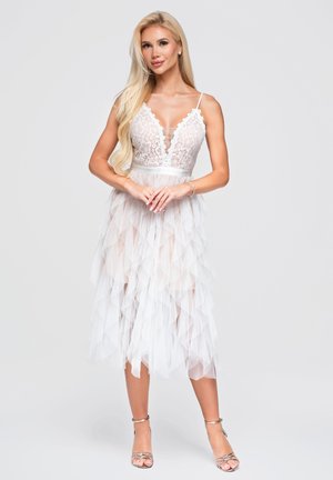 Femme blonde aux longs cheveux portant une robe à corsage en dentelle blanche avec une jupe en tulle superposée et des talons ouverts argentés, se tenant devant un fond uni.