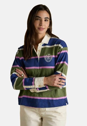 Junge Frau mit langen dunklen Haaren, die ein gestreiftes Rugby-Shirt in Grün, Blau und Pink mit einem weißen Kragen und hellbeigen Hosen trägt, mit verschränkten Armen.