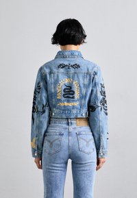 Šviesiai mėlynas džinsinis trumpas švarkas su juodais gėlių siuvinėjimais, geltona "MOSCHINO JEANS" užrašas ir atitinkančios šviesiai mėlynos džinsų kelnės.