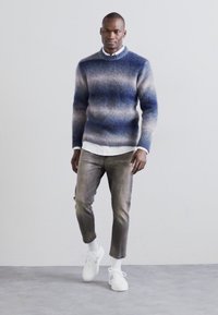 Strikkegenser i gradient blåtoner med ribbestrikket krage, kombinert med lysegrå bukser og hvite sneaks. Avslappet passform, moderne design.
