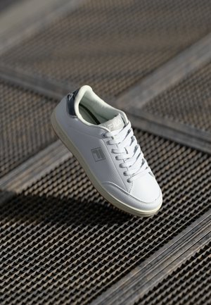 Hvite tennissko med teksturert overflate, gummisåle, snøring og grå detalj på hælen. Fila-logo på siden og tungen.