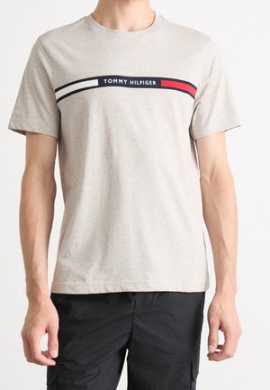 T-shirt gris en coton avec un col rond. Présente une bande noire avec le texte "TOMMY HILFIGER" et des rayures rouges et blanches sur la poitrine.