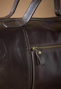 Bolsa de viaje de cuero marrón con textura lisa, cremallera dorada y doble asa. Presenta costuras detalladas y un pequeño bolsillo lateral.