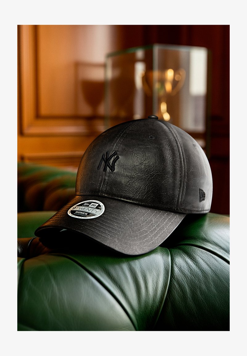 Schwarze Leder-Baseballmütze mit geprägt Logo, strukturierter Krone, gebogenem Schirm und Etikettendetails. Vor grünem Lederhintergrund.