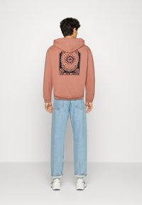 Felpa con cappuccio color rosa polveroso, con grafica nera sul retro; abbinata a jeans dritti di colore blu chiaro. Materiale in cotone.