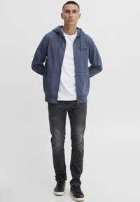 Felpa blu con zip frontale, due tasche laterali e cappuccio con cordino; indossata sopra una maglietta bianca e abbinata a jeans scuri e sneaker nere.