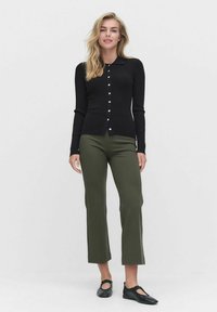 Kvinna som står klädd i en svart knappad kofta, olivgröna cropped byxor och svarta Mary Jane-stil skor mot en neutral bakgrund.