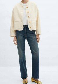 Veste en fausse fourrure ivoire au design court, boutons dorés larges, poches latérales, associée à un jean bleu taille haute et des chaussures dorées.