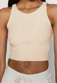 Haut de sport court beige clair avec une texture lisse, un col rond et des coutures décoratives. Présente des motifs en relief subtils sur le devant.