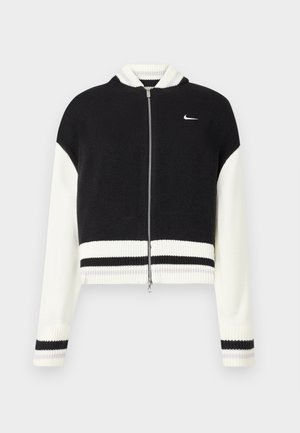 Giacca cropped nera e bianca con zip e orlo a coste. Presenta una texture a maglia e un piccolo logo sul lato sinistro del petto. Accenti a righe sulle maniche.