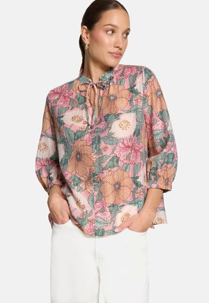 Woman wearing a pink floral blouse with long sleeves and tie neckline, paired with white pants, hands in pockets, looking sideways.