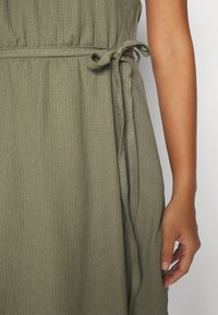 Zeleno-olivové šaty s texturovanou látkou a vázankou v pase. Ruka viditelná na straně, zvýrazňuje střih oděvu.