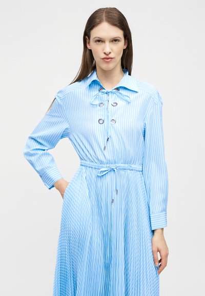 Γυναίκα που φοράει ένα φόρεμα μακρυμάνικο με ριγέ σχέδιο σε ανοιχτό γαλάζιο και λευκό, με γιακά, κορδόνια στο μπροστινό μέρος και δεμένη ζώνη στη μέση, μπροστά σε απλό φόντο.