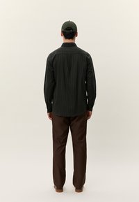 Άνδρας που φοράει σκούρα ριγέ μακρυμάνικη μπλούζα, καφέ παντελόνι και πράσινο καπέλο, στέκεται με την πλάτη προς το μέρος μας μπροστά σε απλό φωτει�νό φόντο.