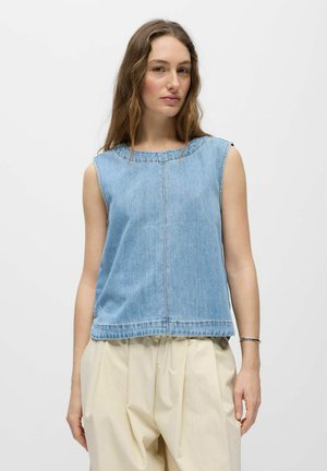 Femme aux cheveux longs et ondulés portant un haut en denim bleu clair sans manches et un pantalon ample beige, debout devant un fond blanc uni.